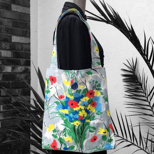 Sac cabas souple pliable fermé par bride en tissu et bouton vintage. Sac fleuri, fleurs stylisées. Au recto le totebag présente une toile de coton texturé (barkcloth), un imprimé de bouquets de fleurs stylisées en rouge bleu jaune vert sur fond blanc et gris. Recto face A.