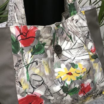 Cabas en toile vintage peinte à la main. Dessin réalisé à grand traits noirs esquissant des oiseaux, des personnages, une femme-oiseau (harpie ?), des insectes, une toile d’araignée, un verre de vin, sur fond abstrait en nuances de gris et touches de rouge, rose, jaune et vert représentant des fleurs. Recto face A, avec bouton, détail.