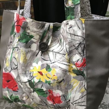 Cabas en toile vintage peinte à la main. Dessin réalisé à grand traits noirs esquissant des oiseaux, des personnages, une femme-oiseau (harpie ?), des insectes, une toile d’araignée, un verre de vin, sur fond abstrait en nuances de gris et touches de rouge, rose, jaune et vert représentant des fleurs. Recto face A, avec bouton, détail et plan serré.