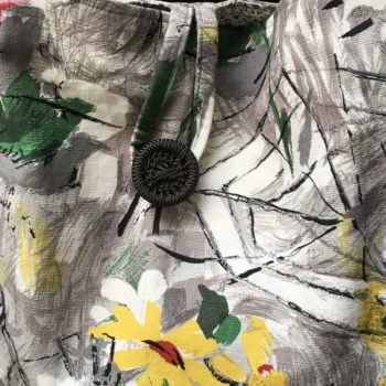 Cabas en toile vintage peinte à la main. Dessin réalisé à grand traits noirs esquissant des oiseaux, des personnages, une femme-oiseau (harpie ?), des insectes, une toile d’araignée, un verre de vin, sur fond abstrait en nuances de gris et touches de rouge, rose, jaune et vert représentant des fleurs. Recto face A, gros plan sur le bouton.