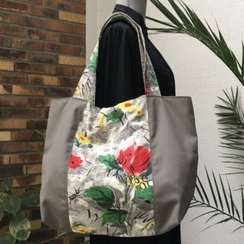 Cabas en toile vintage peinte à la main. Dessin réalisé à grand traits noirs esquissant des oiseaux, des personnages, une femme-oiseau (harpie ?), des insectes, une toile d’araignée, un verre de vin, sur fond abstrait en nuances de gris et touches de rouge, rose, jaune et vert représentant des fleurs. Recto face B.