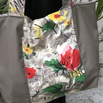 Cabas en toile vintage peinte à la main. Dessin réalisé à grand traits noirs esquissant des oiseaux, des personnages, une femme-oiseau (harpie ?), des insectes, une toile d’araignée, un verre de vin, sur fond abstrait en nuances de gris et touches de rouge, rose, jaune et vert représentant des fleurs. Recto face B, détail.