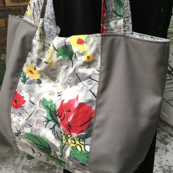 Cabas en toile vintage peinte à la main. Dessin réalisé à grand traits noirs esquissant des oiseaux, des personnages, une femme-oiseau (harpie ?), des insectes, une toile d’araignée, un verre de vin, sur fond abstrait en nuances de gris et touches de rouge, rose, jaune et vert représentant des fleurs. Recto face B, détail autre angle.