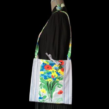 Sacoche mains libres à motif de fleurs. Au recto le sac présente une toile de coton texturé (barkcloth), un imprimé de bouquets de fleurs stylisées en rouge bleu jaune vert sur fond blanc et gris. Porté à l'épaule.