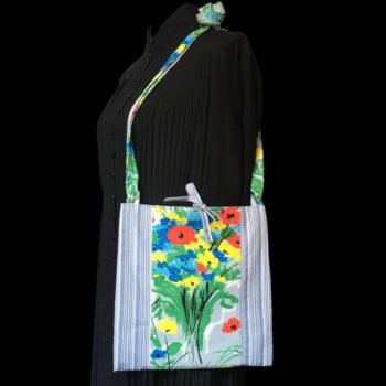 Sacoche mains libres à motif de fleurs. Au recto le sac présente une toile de coton texturé (barkcloth), un imprimé de bouquets de fleurs stylisées en rouge bleu jaune vert sur fond blanc et gris. Recto face B. Porté à l'épaule.