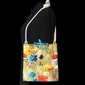 Sac bandoulière à imprimé fleuri, en tissu vintage. Au recto le sac présente une toile en coton texturé des années 1960s, au dessin de fleurs des champs en bleu jaune orange blanc sur fond vert. Recto face B. Porté à l'épaule.