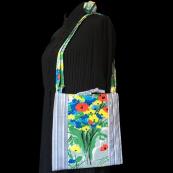 Sacoche mains libres à motif de fleurs. Au recto le sac présente une toile de coton texturé (barkcloth), un imprimé de bouquets de fleurs stylisées en rouge bleu jaune vert sur fond blanc et gris. Recto face A. Porté à l'épaule.