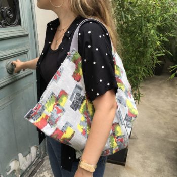 Cabas triangle en pliage origami, coupon vintage des années 1950s début 1960s, tissu souple avec effet texturé et satiné. Imprimé graphique, motifs géométriques abstraits époque moderne atomique milieu XXe siècle, dessins en noir rouge jaune vert gris sur fond gris clair. Vu porté, de profil.