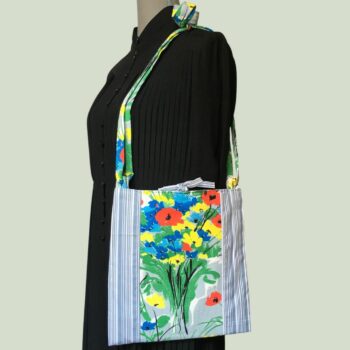 Sacoche mains libres à motif de fleurs. Au recto le sac présente une toile de coton texturé (barkcloth), un imprimé de bouquets de fleurs stylisées en rouge bleu jaune vert sur fond blanc et gris. Recto face A. Porté à l'épaule.
