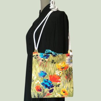 Sac bandoulière à imprimé fleuri, en tissu vintage. Au recto le sac présente une toile en coton texturé des années 1960s, au dessin de fleurs des champs en bleu jaune orange blanc sur fond vert. Recto face A. Porté à l'épaule.