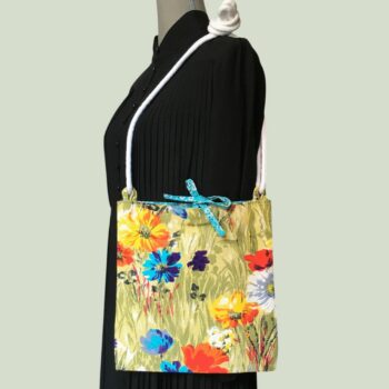 Sac bandoulière à imprimé fleuri, en tissu vintage. Au recto le sac présente une toile en coton texturé des années 1960s, au dessin de fleurs des champs en bleu jaune orange blanc sur fond vert. Recto face B. Porté à l'épaule.