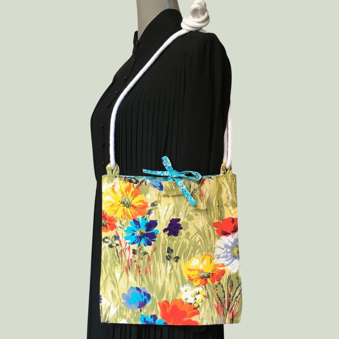 Sac bandoulière à imprimé fleuri, en tissu vintage. Au recto le sac présente une toile en coton texturé des années 1960s, au dessin de fleurs des champs en bleu jaune orange blanc sur fond vert. Recto face B. Porté à l'épaule.