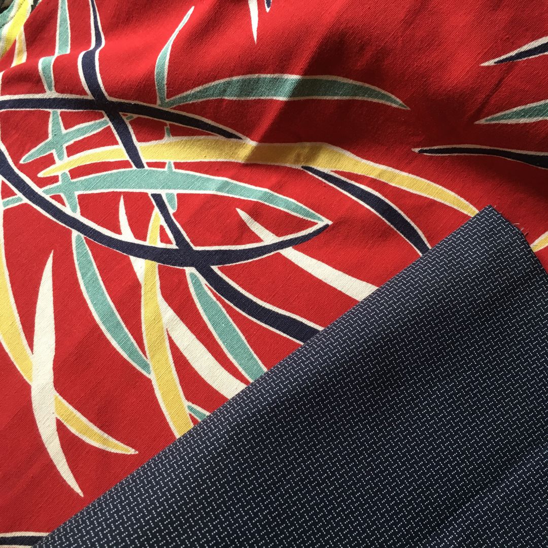 Le tissu Gai Color, toile d’ameublement vintage typique des années 1950s, en coton. L’imprimé présente un motif tropical et exotique d’inspiration hawaïenne. Sur un fond rouge vif, de grandes feuilles stylisées (rappelant des frondes de palmier ou des fougères) s’entrelacent. Les feuilles utilisent des teintes contrastées de turquoise / vert d’eau, de jaune pastel et de bleu marine très foncé. Les lignes de contour blanches soulignent le dynamisme et le mouvement fluide du motif.