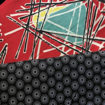 Lorraine Fleurie, toile d’ameublement vintage typique des années 1950s, en coton. Le fond rouge vif saturé sert de base éclatante à l'ensemble du motif. Des triangles ou trapèzes inversés de couleur turquoise (bleu canard) sont surmontés ou soulignés d'une touche de jaune. Une multitude de lignes droites et fines, noires et blanches, s'entrecroisent de manière chaotique et dynamique par-dessus et autour des formes colorées asymétriques. Ce réseau de lignes évoque des structures filaires, des baguettes ou des constellations géométriques. Ici avec le tissu d'accompagnement (doublure).
