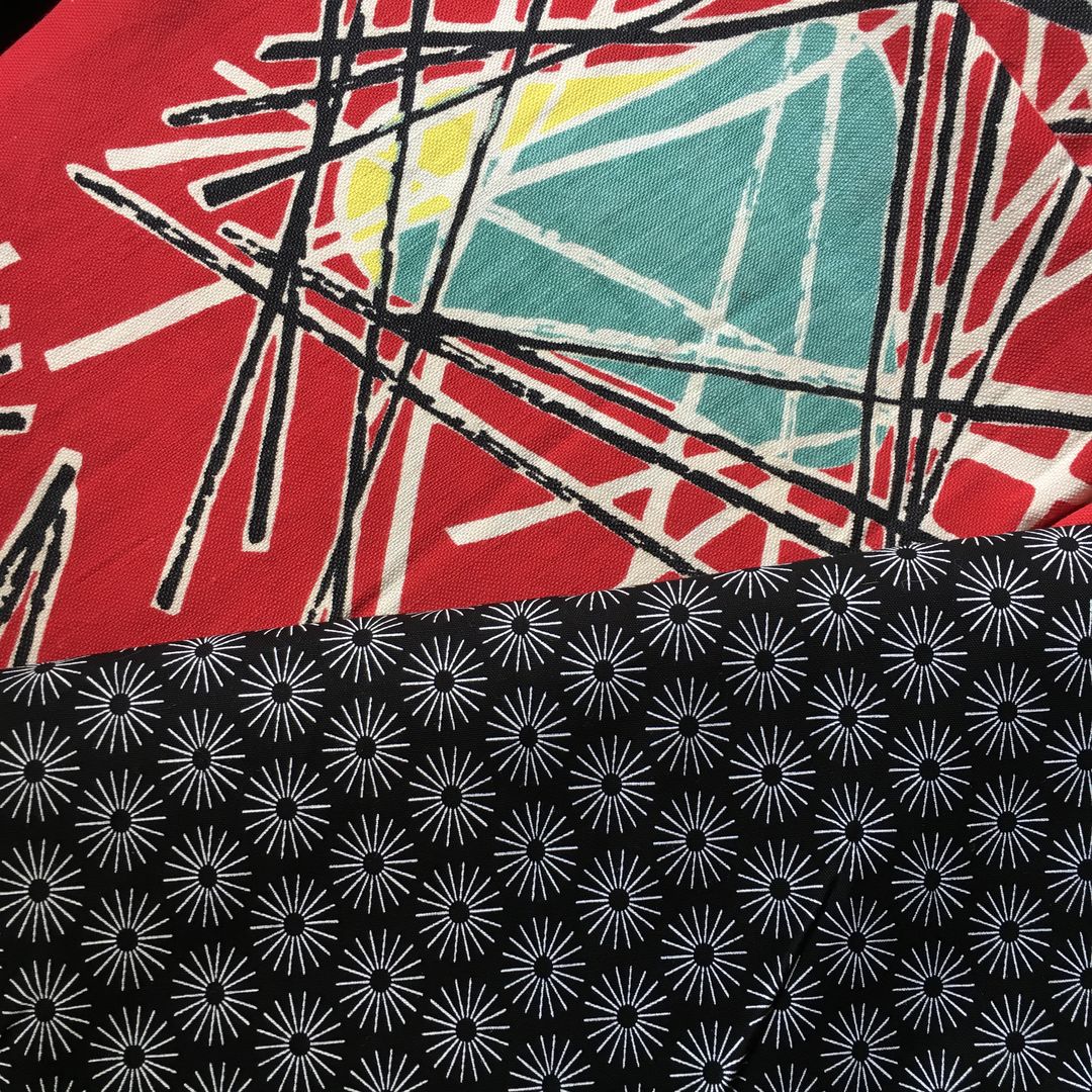 Lorraine Fleurie, toile d’ameublement vintage typique des années 1950s, en coton. Le fond rouge vif saturé sert de base éclatante à l'ensemble du motif. Des triangles ou trapèzes inversés de couleur turquoise (bleu canard) sont surmontés ou soulignés d'une touche de jaune. Une multitude de lignes droites et fines, noires et blanches, s'entrecroisent de manière chaotique et dynamique par-dessus et autour des formes colorées asymétriques. Ce réseau de lignes évoque des structures filaires, des baguettes ou des constellations géométriques. Ici avec le tissu d'accompagnement (doublure).