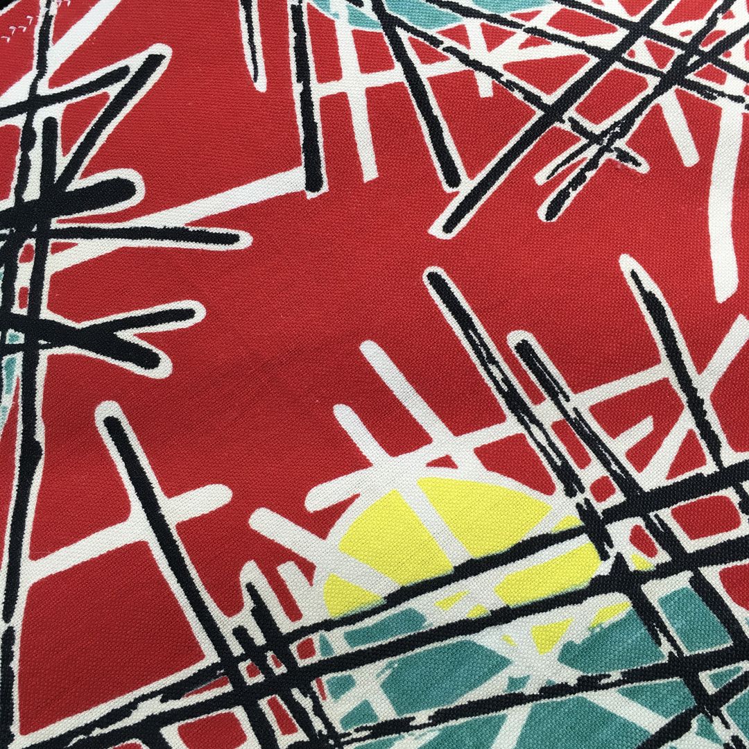 Lorraine Fleurie, toile d’ameublement vintage typique des années 1950s, en coton. Le fond rouge vif saturé sert de base éclatante à l'ensemble du motif. Des triangles ou trapèzes inversés de couleur turquoise (bleu canard) sont surmontés ou soulignés d'une touche de jaune. Une multitude de lignes droites et fines, noires et blanches, s'entrecroisent de manière chaotique et dynamique par-dessus et autour des formes colorées asymétriques. Ce réseau de lignes évoque des structures filaires, des baguettes ou des constellations géométriques.